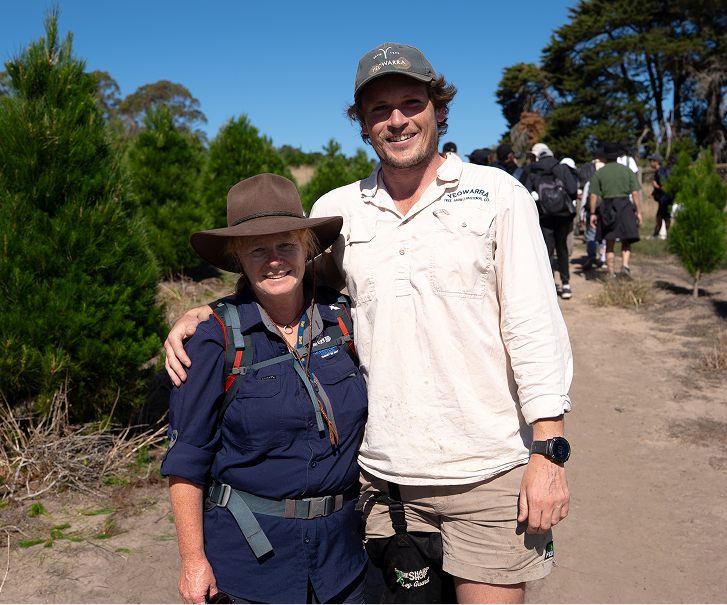A happy photo of a school teacher with Yeowarra tour guide, Josh. In the background there are large pine trees and a group of school students.