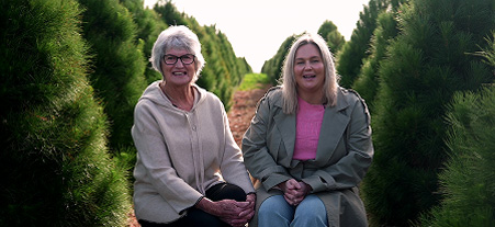 A photo of a grandmother and her daughter, sitting in a row of pine trees facing the camera.